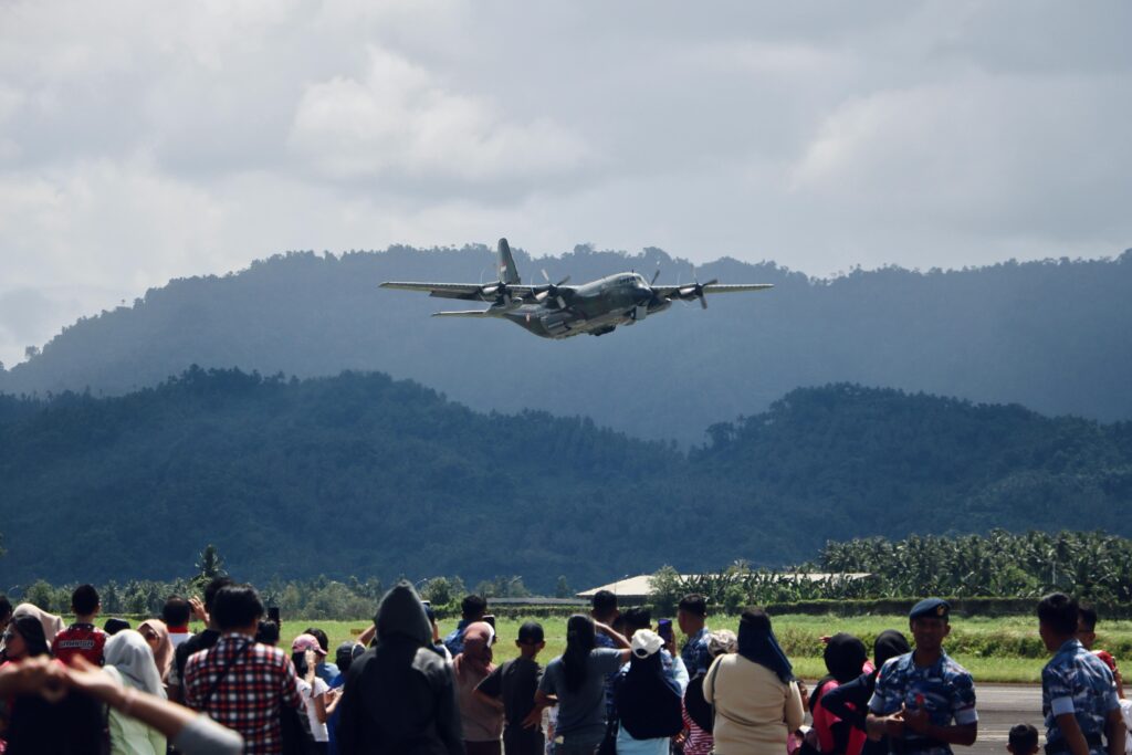A military plane takes off in Sulawesi, Indonesia observed by a crowd with lush mountains in the background.