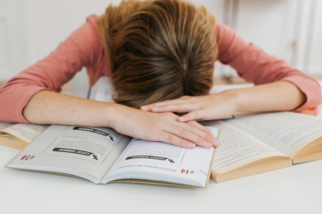 A student overwhelmed by studying falls asleep on open books at a desk, representing academic fatigue.