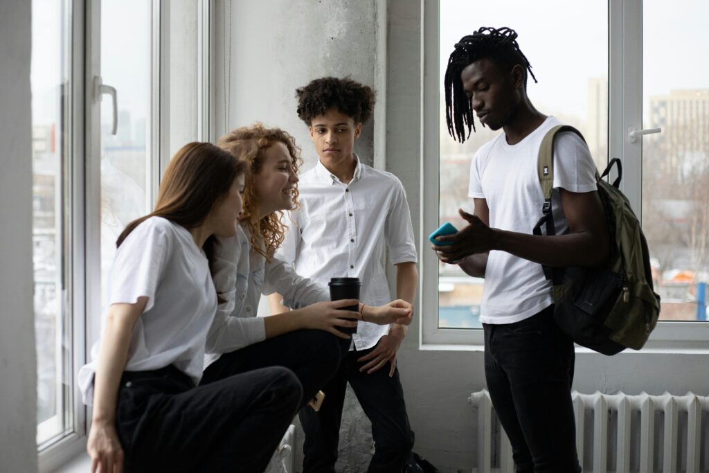 Group of multiracial classmates with backpack and takeaway coffee having conversation at windowsill near window in light classroom before lesson