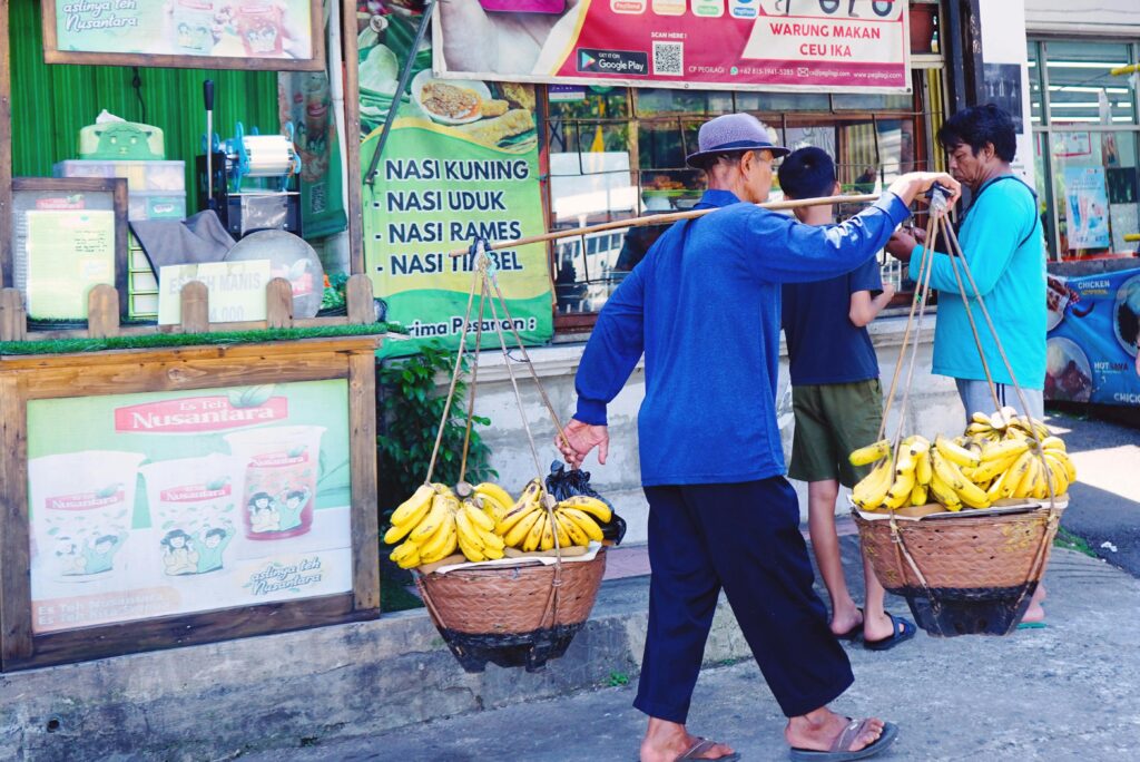 A street vendor carrying bananas in Bandung, Indonesia, near a local food stall. Urban daily life scene.