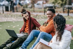 Group of multiracial students sitting on grassy ground with book browsing laptop while studying together on street against blurred background
