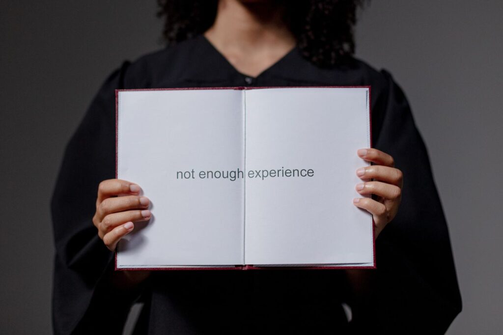 A graduate in cap and gown displays a diploma reading 'not enough experience', highlighting job market challenges.