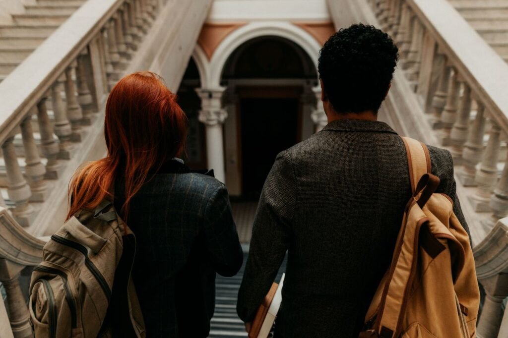 Two university students with backpacks walk down a grand staircase in a historic building.