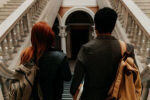 Two university students with backpacks walk down a grand staircase in a historic building.